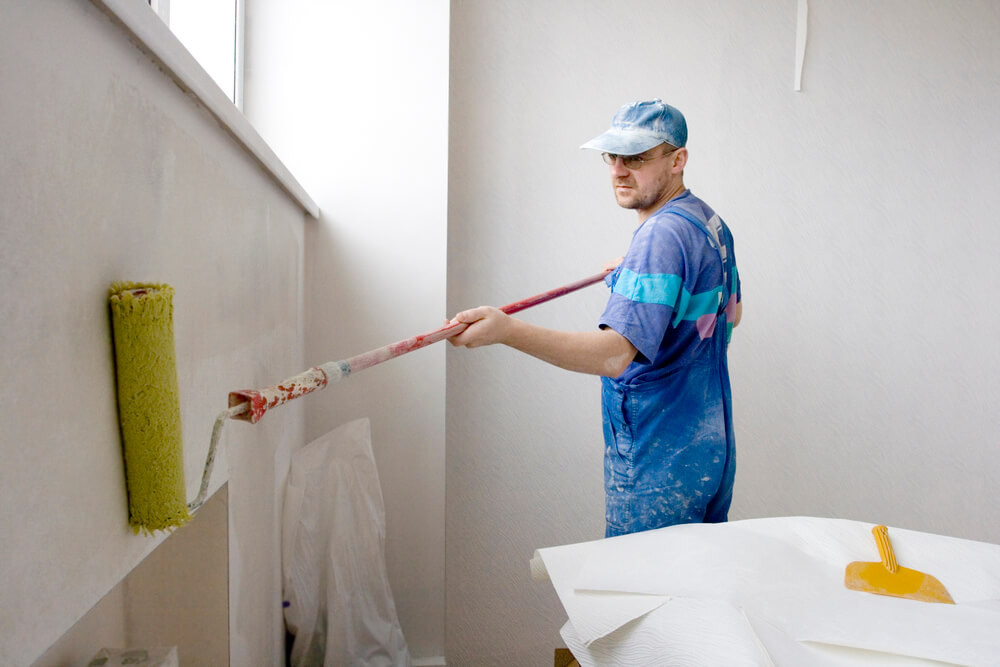 One of our shop painters with a roller brush painting a white wall whilst wearing a painting cap and overalls