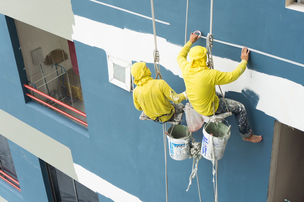two external painters on a harness with a paint bucked hanging below and painting the outside of a blue building